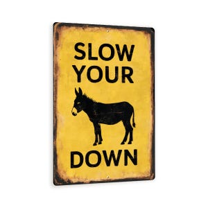May include: A rectangular metal sign with a yellow background and black text and a black donkey silhouette. The text reads "SLOW YOUR DOWN" in a bold, sans-serif font. The sign has a distressed, vintage look with a dark brown border.