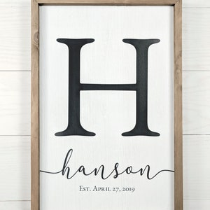 A white wood family established sign with monogram hanging on a shiplap wall. The sign has an H for a monogram and displays the family surname in an attractive script font.  the established date is shown in a stately serif font.