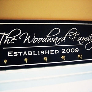 May include: Black wooden sign with white lettering that reads "The Woodward Family Established 2009". The sign has five gold hooks.