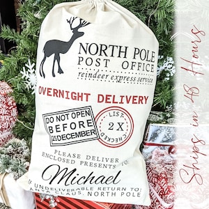 May include: A white canvas Santa sack with a black reindeer illustration and the text "NORTH POLE POST OFFICE reindeer express service OVERNIGHT DELIVERY DO NOT OPEN BEFORE 25 DECEMBER LIST 2 X CHECKED PLEASE DELIVER TO: Michael IF UNDELIVERABLE RETURN TO: SANTA CLAUS. NORTH POLE"