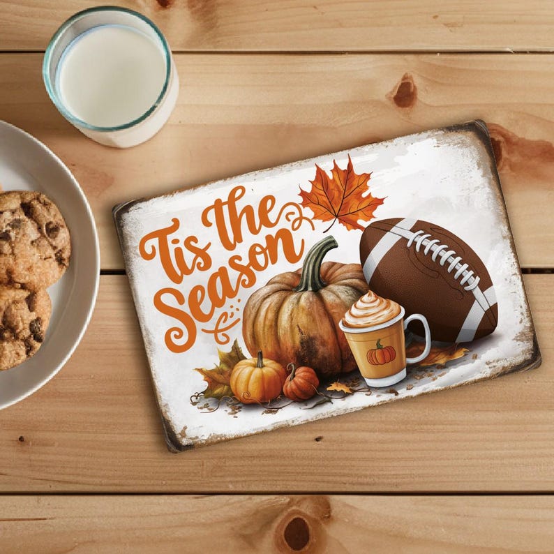 May include: White sign with the text "Tis the Season" in orange lettering. The sign features a brown football, a pumpkin, a cup of coffee with whipped cream, and a small pumpkin. There are also fall leaves and acorns on the sign.