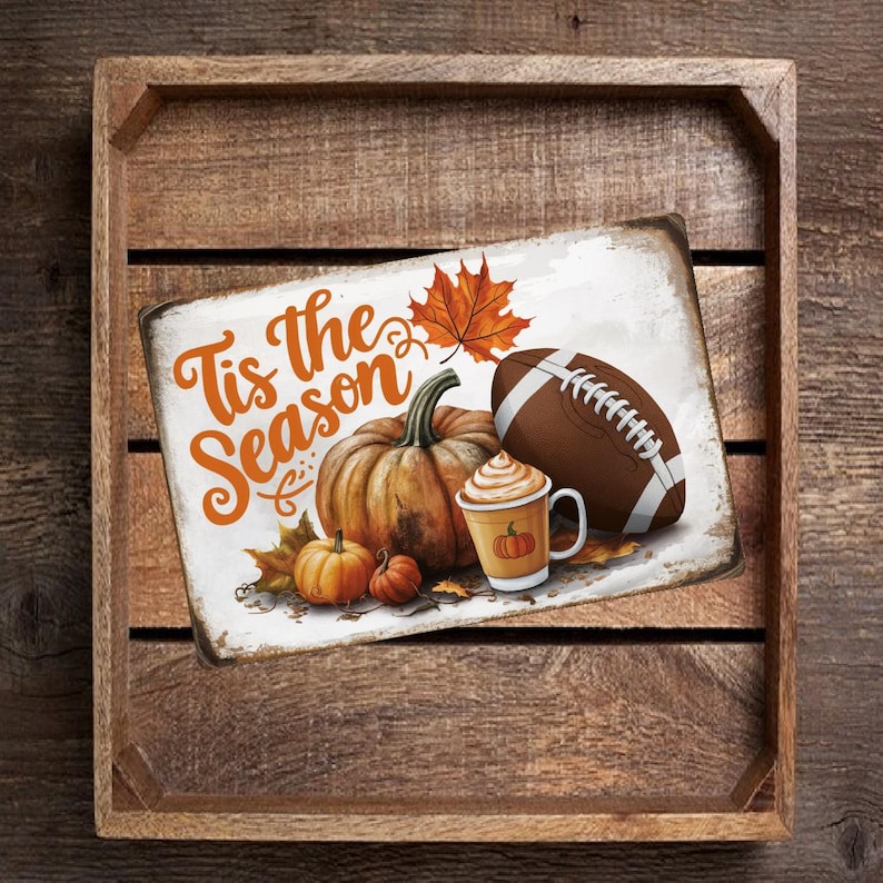 May include: A white metal sign with the text "Tis the Season" in orange and brown lettering. The sign also features a football, a pumpkin, a cup of coffee with whipped cream, and fall leaves.