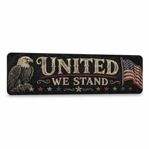 May include: A rectangular black sign featuring a white bald eagle on the left and an American flag on the right. The text UNITED WE STAND is printed in a white, stylized font in the center, accented by a row of small red, white, and blue stars.