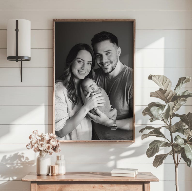 May include: Black and white photograph of a family of three. A man and woman are holding a newborn baby wrapped in a blanket. The photo is framed in a simple wooden frame.