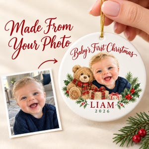 Puede incluir: Un adorno navideño redondo de cerámica blanca con la foto de un bebé sonriente, un oso de peluche con bufanda de cuadros y regalos. El texto dice Baby's First Christmas, LIAM y 2026, rodeado de ramas de pino verde y bayas rojas.