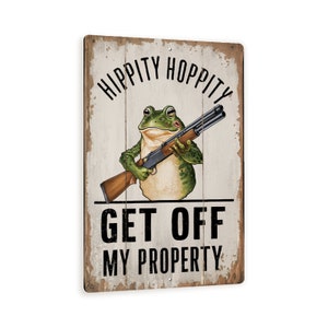 May include: A rustic wooden sign with a cartoon frog holding a shotgun. The sign reads "Hippity Hoppity Get Off My Property".