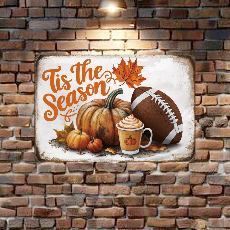 May include: A metal sign with the text "Tis the Season" in orange and white lettering. The sign features a brown football, a pumpkin, and a cup of coffee with whipped cream and a small pumpkin on top. The sign is decorated with fall leaves and pumpkins.
