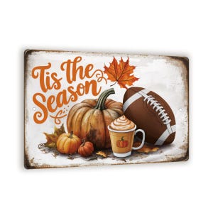 May include: A white sign with the text "Tis the Season" in orange lettering. The sign features a football, a pumpkin, and a cup of pumpkin spice latte. There are also fall leaves and smaller pumpkins around the main objects.