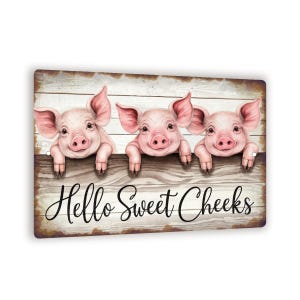 May include: A wooden sign featuring three pink pigs peeking over a wooden fence. The pigs have large ears and are smiling. The sign has the text "Hello Sweet Cheeks" in a cursive font.