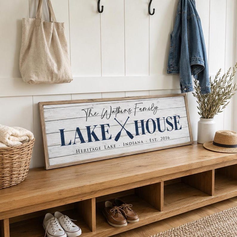 May include: A wooden sign with the text "The Watkins Family LAKE HOUSE" in navy blue, with crossed oars. The sign is on a wooden bench with shoes and a basket. A beige tote bag hangs on the wall.