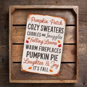May include: A white metal sign with a rustic look featuring the text "Pumpkin Patch Cozy Sweaters Cuddles and Snuggles Falling Leaves Warm Fireplaces Pumpkin Pie Laughter & Joy It's Fall". The sign has a red and orange fall leaf design.