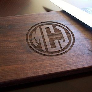 May include: A dark brown wooden cutting board with a handle and a circular monogram of the letters "MCJ" engraved in the center. A silver knife is lying on the board.