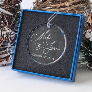 May include: A clear glass ornament with a blue box. The ornament is engraved with the text "Mike & Jenni October 3rd, 2023".