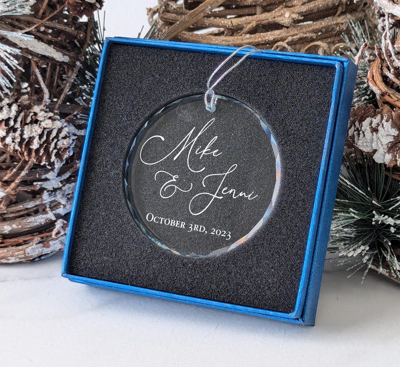 May include: A clear glass ornament with a blue box. The ornament is engraved with the text "Mike & Jenni October 3rd, 2023".