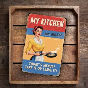 Puede incluir: Un letrero de metal de estilo vintage con un esquema de color rojo, amarillo y azul. Muestra a una mujer con un vestido amarillo y un delantal azul cocinando huevos en una sartén. El texto en el letrero dice "My Kitchen My Rules! Today's Menu?? Take it or Leave it!"