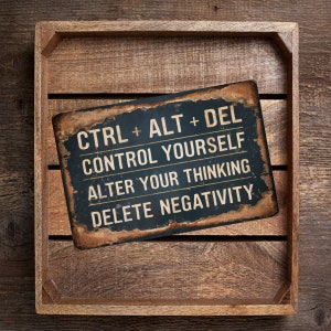 May include: A rectangular, distressed metal sign with the text "CTRL + ALT + DEL CONTROL YOURSELF ALTER YOUR THINKING DELETE NEGATIVITY" in white lettering. The sign is set in a wooden tray.