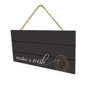 May include: Black wooden sign with a white rope hanger. The sign has a distressed wood look and says "make a wish" in white script with a gold swirl design.