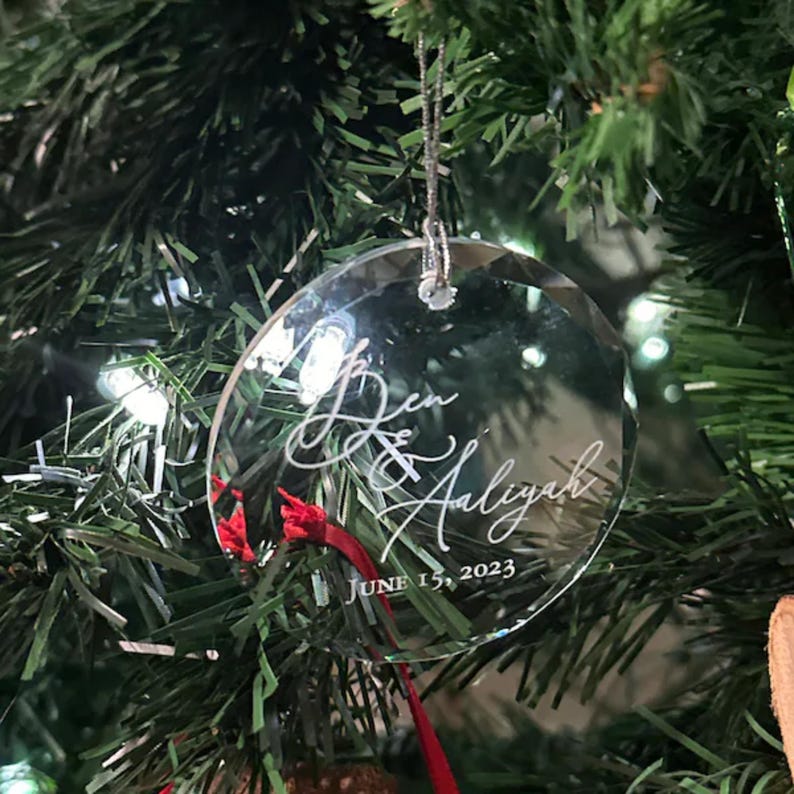 May include: A clear glass ornament with the names "Ben & Aliyah" and the date "June 15, 2023" etched on it. The ornament is hanging on a Christmas tree.