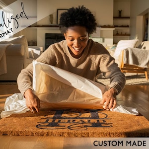 A woman is opening gift of a personalized coir door mat. There is gift wrapping on the floor and she is smiling excitedly at receiving the gift.  There is a large monogram initial printed on the mat along with the recipients last name.