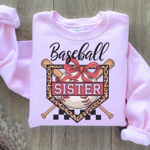 Puede incluir: Sudadera rosa con un gráfico de béisbol. El gráfico presenta una pelota de béisbol en un plato de home con bates cruzados detrás. La pelota de béisbol tiene un lazo rojo y el texto "Baseball Sister" está en el plato de home.