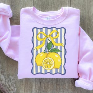 May include: A pink sweatshirt with a graphic of three lemons with a yellow bow on a blue and white striped background.