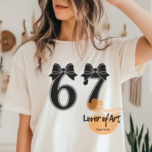 May include: Cream-colored t-shirt featuring the number "67" in black, adorned with black bows and a decorative border. Below the numbers, the text "Lover of Art" and "Design Studio" are printed in black.