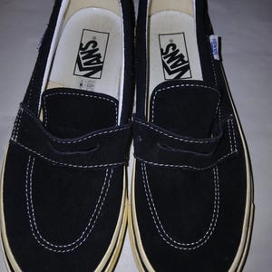May include: Pair of black Vans slip-on shoes with white stitching and a cream-colored sole. The shoes feature the Vans logo on the insole and a small tag on the side. The shoes are made of suede material.