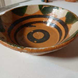 May include: A brown ceramic bowl with a white rim and green and black painted designs. The bowl has a circular pattern in the center.