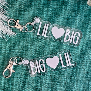 May include: Two clear acrylic keychains with silver-coloured hardware. Each keychain has the words "LIL" and "BIG" with a white heart in between. The keychains are on a textured teal background with white feather and leaf accents.