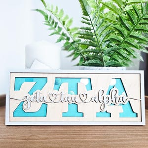 May include: Wooden sign featuring the Greek letters ZTA, with the words "zeta tau alpha" in cursive. The background is light blue. The sign is in a white frame and rests on a wooden surface.