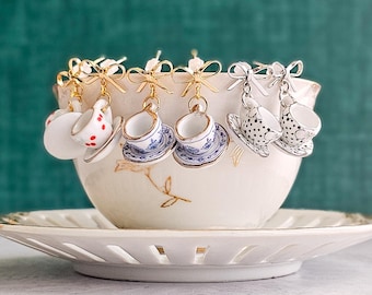 May include: Miniature teacup earrings hanging from gold-coloured bows. The earrings showcase diverse designs, including polka dots and blue floral patterns. They are presented within a white teacup and saucer, evoking a sense of elegance.