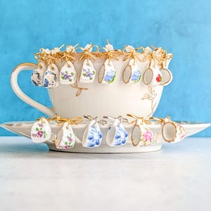 May include: A cream-coloured teacup and saucer set with gold edging, decorated with miniature porcelain teacups. Each small cup has a different floral pattern and is suspended by gold-coloured wire. The background is a solid light blue.