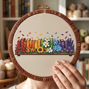 以下が含まれることがあります： カラフルな本棚のデザインが施されたクロスステッチ刺繍枠。本は虹色に配置され、花と葉が添えられています。木製の枠には、装飾的な花のボーダーがあります。
