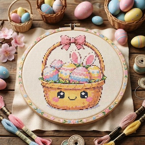 Puede incluir: Un bastidor de bordado de punto de cruz con una cesta de huevos de Pascua con orejas de conejo. La cesta tiene una cara sonriente y un lazo rosa. El bastidor está rodeado de cestas de huevos, hilo de bordar y flores.