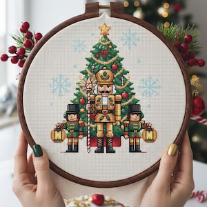 May include: A cross-stitch embroidery hoop depicting a Christmas scene with a tree and nutcracker figures. The tree is adorned with decorations, and the nutcrackers wear gold and green uniforms. The hoop is held by hands with green and gold nail polish.