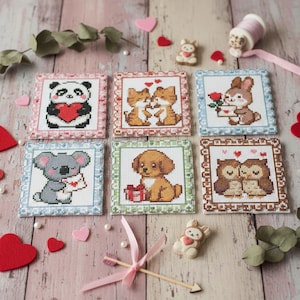 May include: Six square cross-stitch designs showcasing animals like a panda, foxes, a rabbit, a koala, a dog, and owls. Each design has a different coloured border and features the animals with hearts or gifts. The background is light wood.