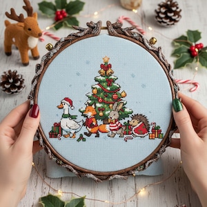 Könnte beinhalten: Ein Kreuzstich-Stickrahmen mit einer Weihnachtsszene. Das Design zeigt einen geschmückten Baum und Tiere in festlicher Kleidung. Der Rahmen hat einen braunen, zweigartigen Rand. Der Hintergrund ist ein hellblauer Stoff.