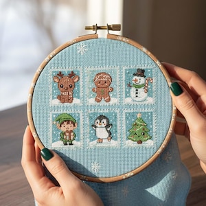 Könnte beinhalten: Ein Kreuzstich-Stickrahmen mit sechs weihnachtlichen Motiven: ein Rentier, ein Lebkuchenmann, ein Schneemann, ein Elf, ein Pinguin und ein Weihnachtsbaum. Die Motive befinden sich auf hellblauem Stoff mit Schneeflocken.