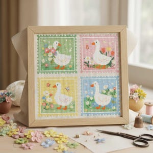 May include: A framed cross-stitch artwork featuring four squares, each with a white goose and floral elements. The squares have different pastel backgrounds: green, pink, yellow, and blue. The frame is made of light-colored wood.