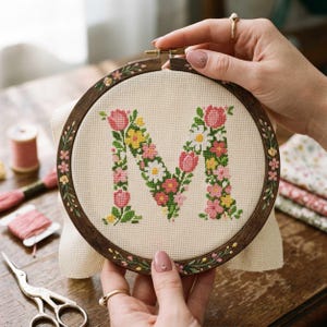 May include: A cross-stitch embroidery hoop with the letter "M" formed from flowers in pink, yellow, and white. The wooden hoop has painted floral details. The embroidery is on cream fabric, with threads and scissors visible on a wooden surface.