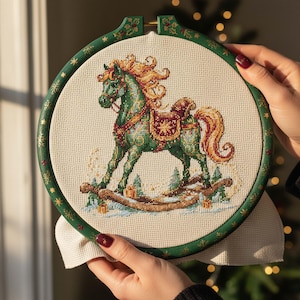 Może przedstawiać: Tamborek do haftu z zielonym wzorem konia na biegunach. Koń ma pomarańczową grzywę i ogon, z ozdobnym siodłem. Zielony tamborek jest ozdobiony złotymi gwiazdami i świątecznymi akcentami, tworząc świąteczny motyw.