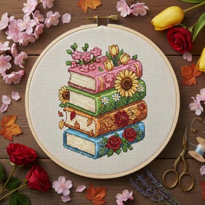 May include: A cross-stitch embroidery hoop featuring a stack of colorful books adorned with flowers. The books are pink, green, orange, and blue, with floral and leaf details. The background is a light-colored fabric, and the hoop is made of wood.