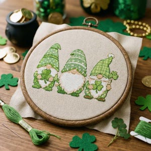 以下が含まれることがあります： 三人の緑色のノームがクローバーを持っているクロスステッチ刺繍。ノームは白いひげと模様入りの帽子をかぶっています。ベージュの布に刺繍されており、緑色の糸とフェルトのクローバーが周りに散らばっています。
