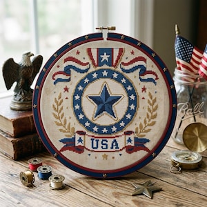 May include: A patriotic cross-stitch embroidery in a round frame, featuring a large blue star at the center, surrounded by a ring of stars and the word "USA" on a banner. The design incorporates red, white, and blue elements, with a dark blue frame. The embroidery is displayed near a window.