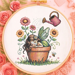Op de afbeelding: Kruissteekborduurwerk met een slapende fee in een bloempot, omringd door bloemen en een gieter. De fee heeft bruin haar en groene vleugels. De borduurwerk zit in een houten hoepel, met roze rozen op de achtergrond.