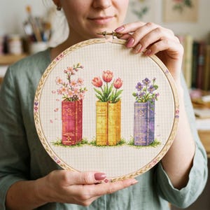 May include: Cross-stitch embroidery in a wooden hoop. The design features three stylized books in shades of pink, yellow, and purple, each holding a different type of flower. The flowers include pink blossoms, red tulips, and purple flowers.