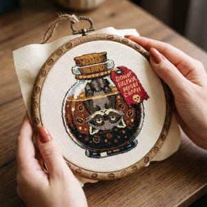 Puede incluir: Bordado de punto de cruz en un aro de madera que representa un mapache dentro de un tarro de cristal con tapa de corcho. El tarro tiene una etiqueta roja que dice "DON'T RELEASE BEFORE COFFEE" con una calavera. El bordado es en tonos marrones, beige y negro.