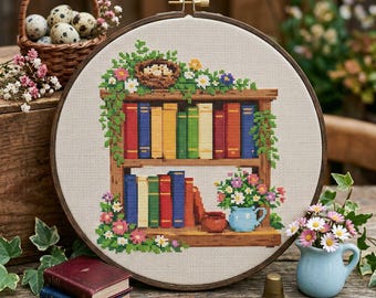 May include: A cross-stitch embroidery hoop depicting a bookshelf with vibrant books, flowers, and foliage. The embroidery is set on a neutral fabric, framed by a wooden hoop. Other items include a basket of eggs, small books, and a blue vase.