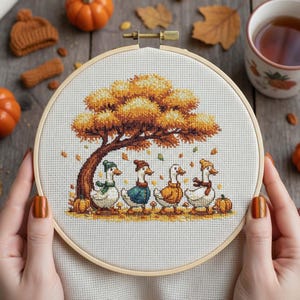 May include: A cross-stitch embroidery hoop featuring a fall scene with four geese under a tree with orange and brown leaves. The geese are wearing hats and scarves. Pumpkins and a small knitted hat are in the background.