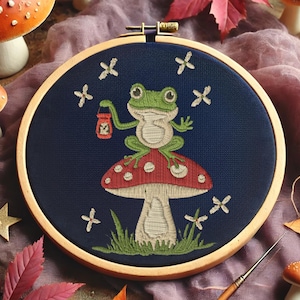May include: An embroidered hoop art piece featuring a green frog sitting atop a red mushroom with white spots. The frog holds a lantern, surrounded by white floral accents on a navy blue background. The wooden hoop adds a rustic touch.